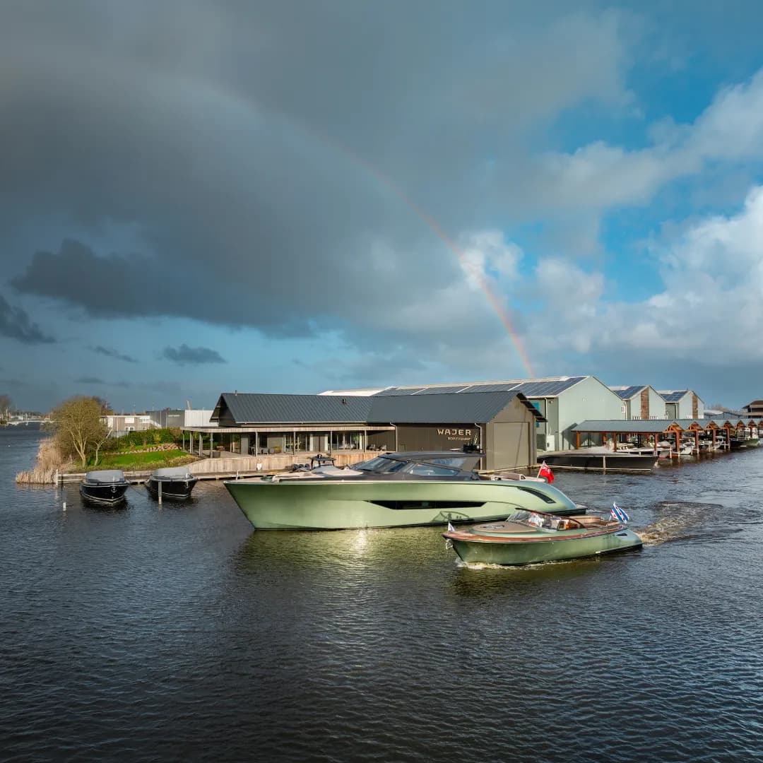 Boats moored on a calm waterway next to modern buildings, with a vibrant rainbow arcing across a partly cloudy sky.