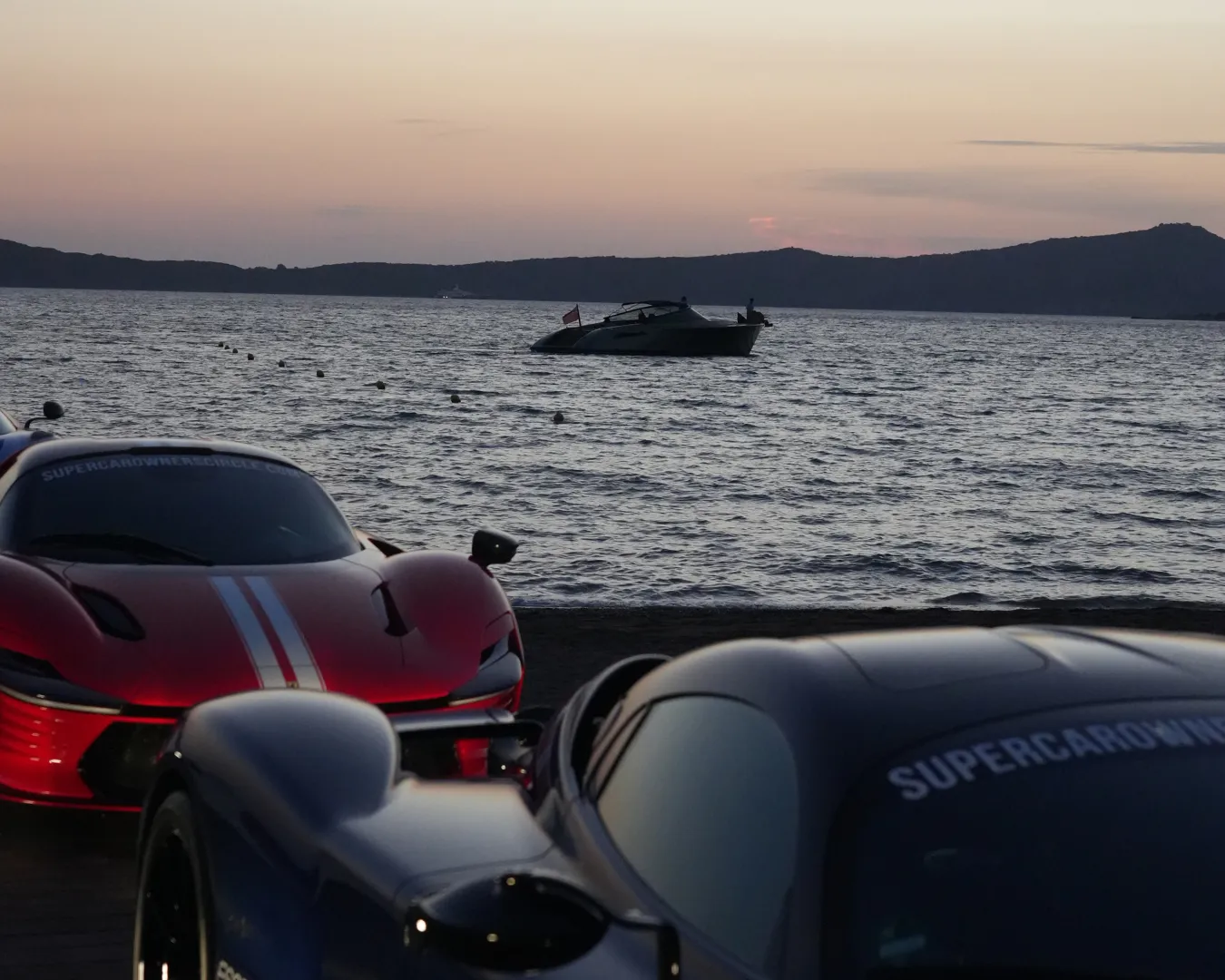 Two sleek cars parked near a beach at sunset, with a boat floating on the calm sea in the background.