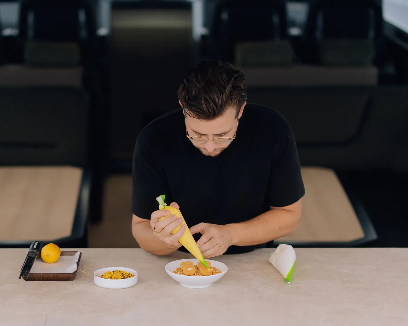 A man in a black shirt decorates a dish with a yellow piping bag in a modern kitchen setting. Bowls of ingredients and a lemon are beside him.