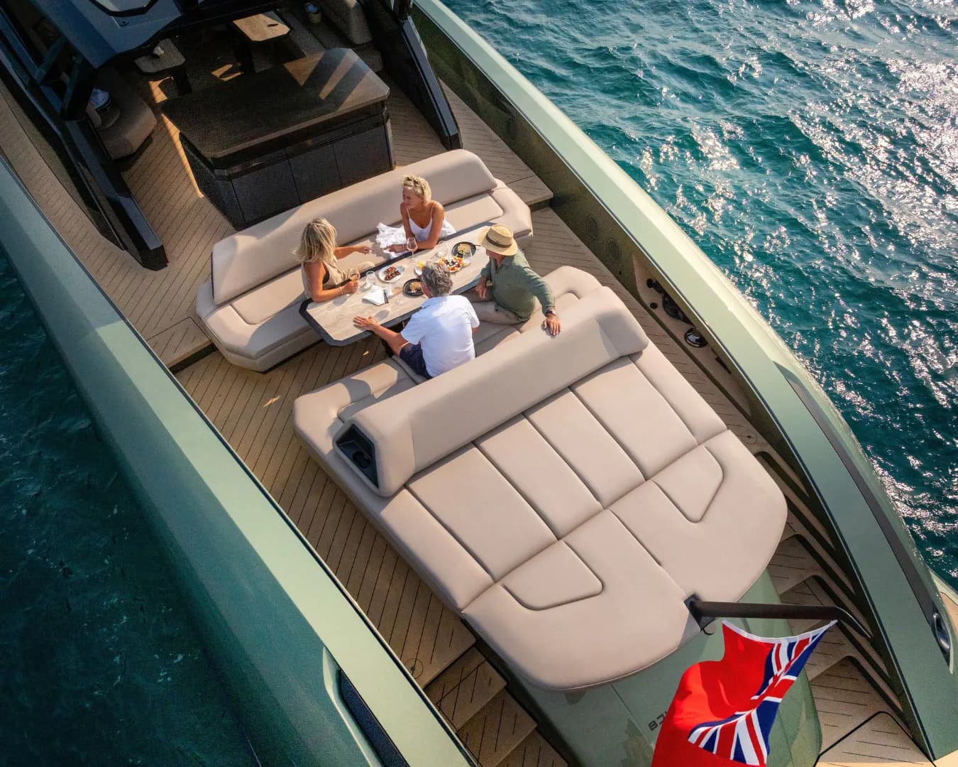 Aerial view of four people enjoying a meal on the deck of a yacht with a British flag, surrounded by clear blue water.