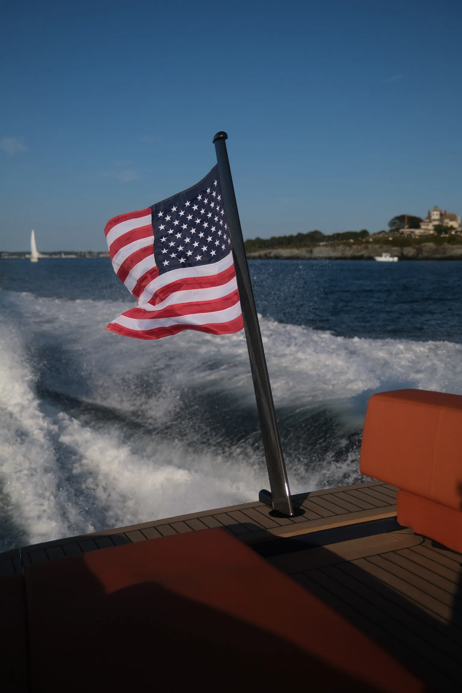 American flag waves on a boat cruising through water, with coastal buildings and a sailboat visible in the background.