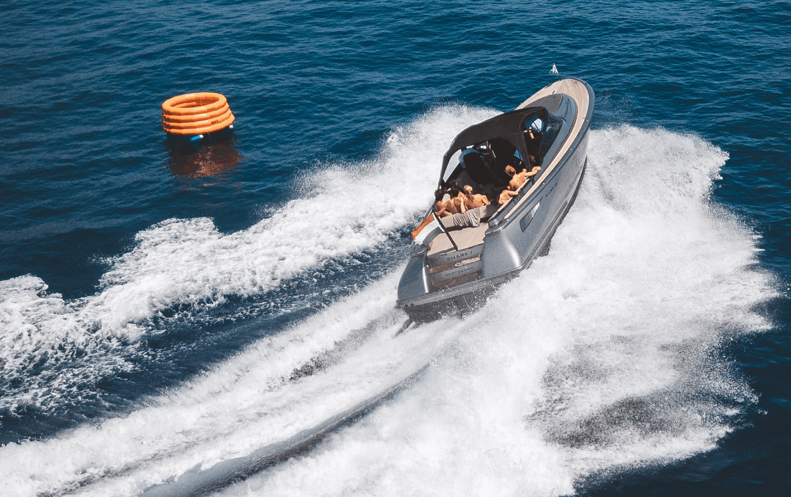 A speedboat with people onboard creates a wake as it moves through the blue ocean, passing an orange inflatable ring floating nearby.