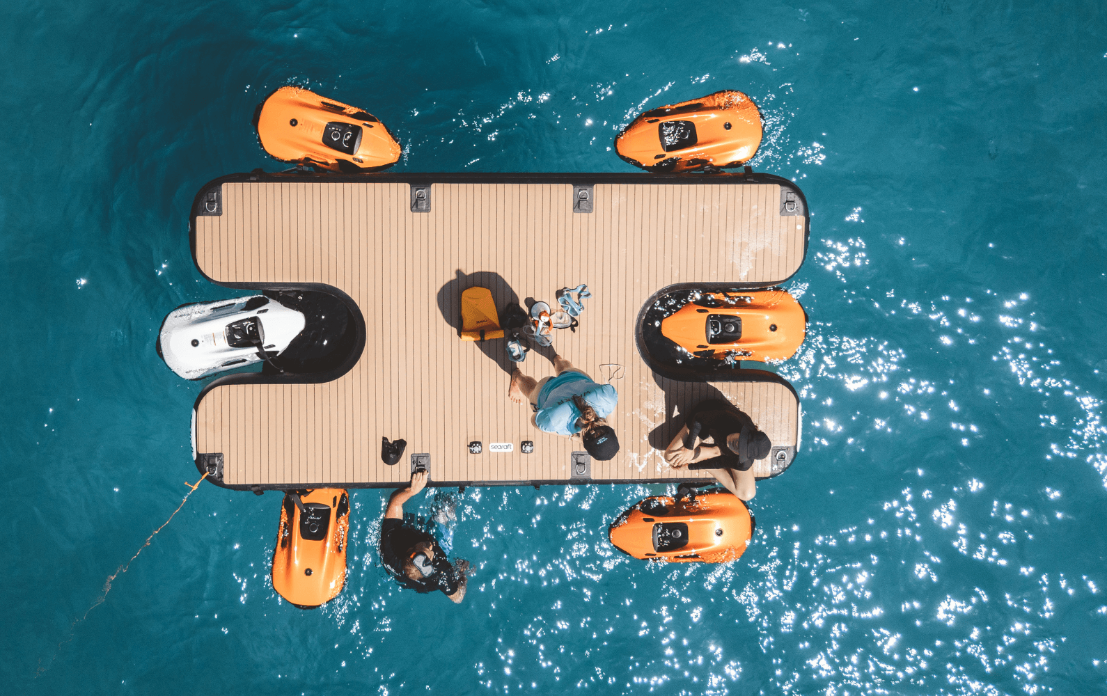 Aerial view of a floating dock with three people and four orange sea scooters in clear blue water, surrounded by sunlight reflections.