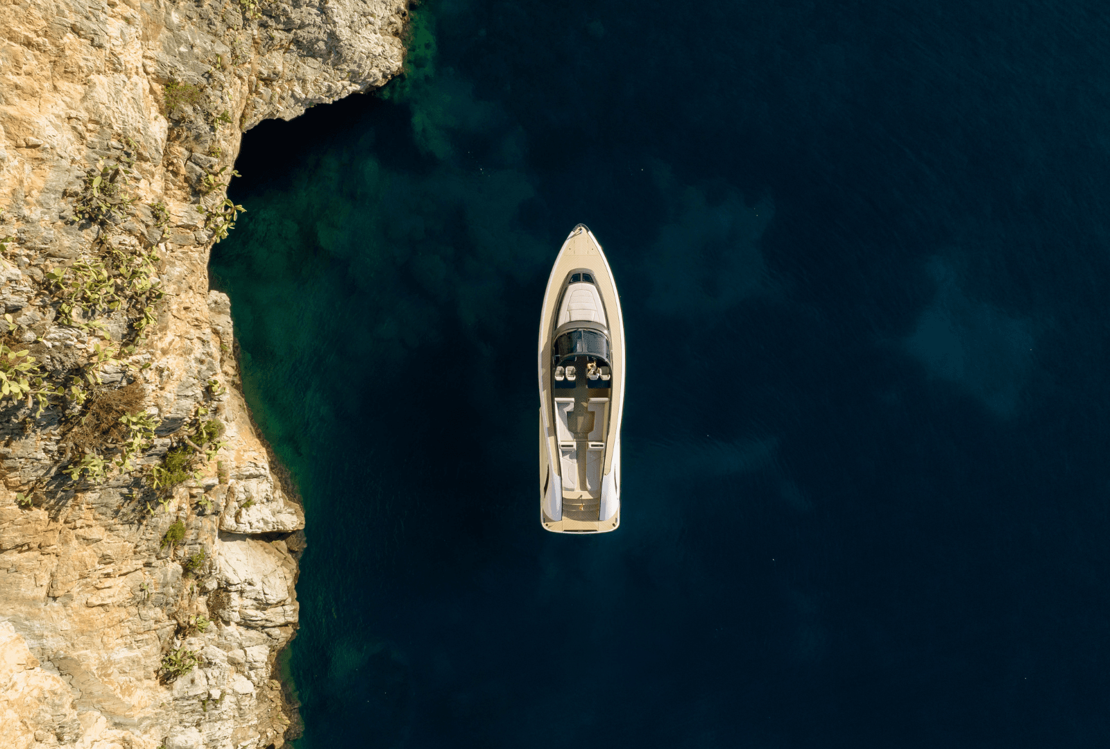 Aerial view of a yacht near a rocky coastline with deep blue water, highlighting the contrast between the natural and man-made elements.