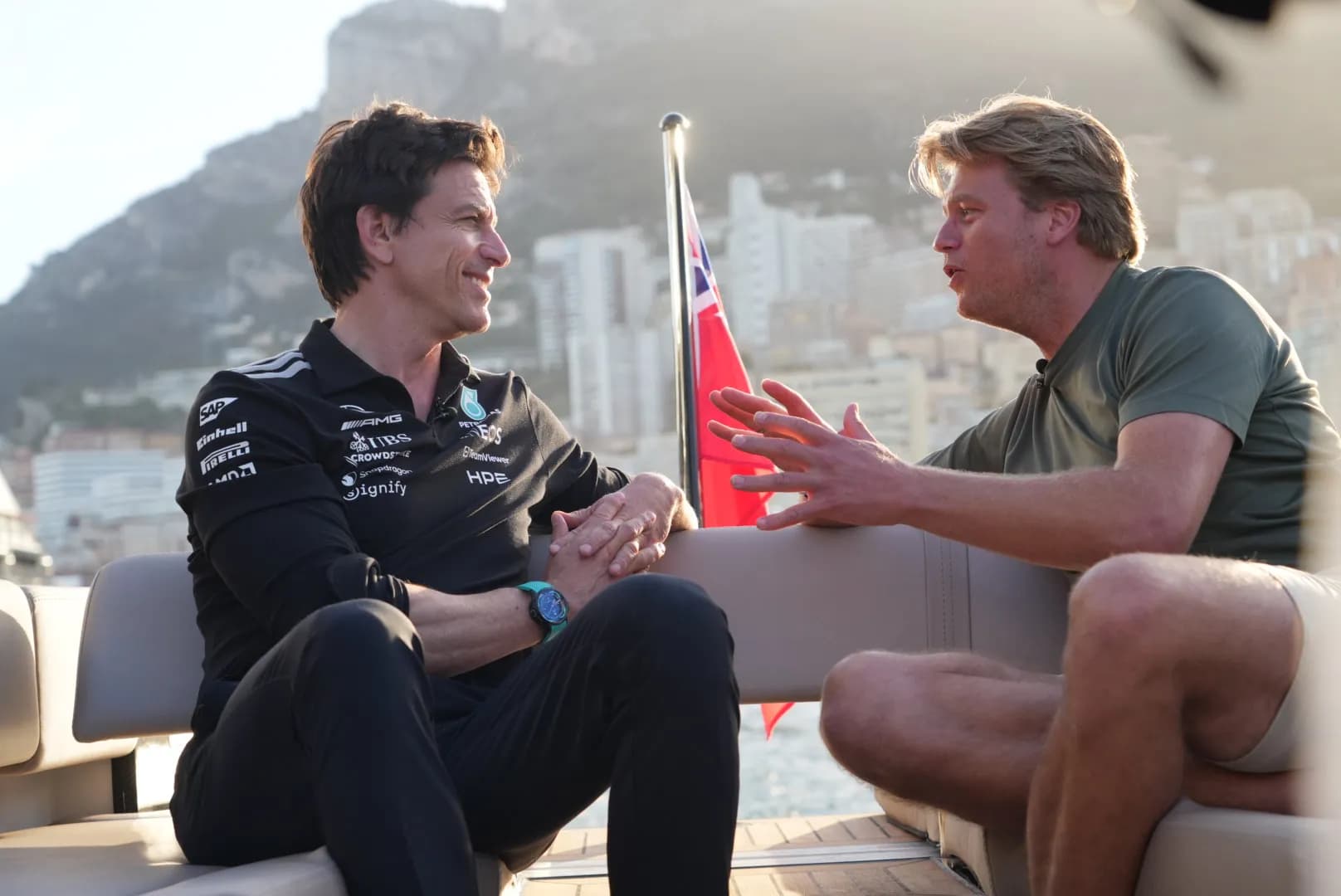 Two men engaged in a lively conversation on a boat, with a cityscape and mountain in the background.