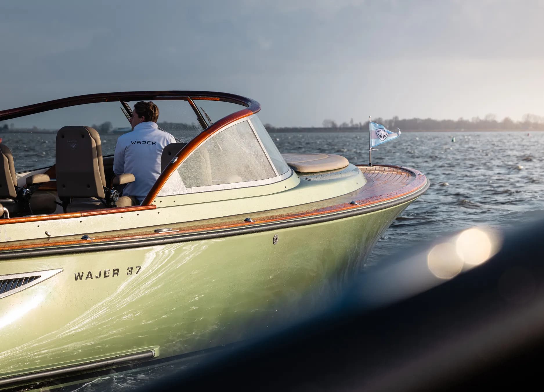 Person in a blue jacket steers a green Wajer 37 boat on a calm sea under cloudy skies.
