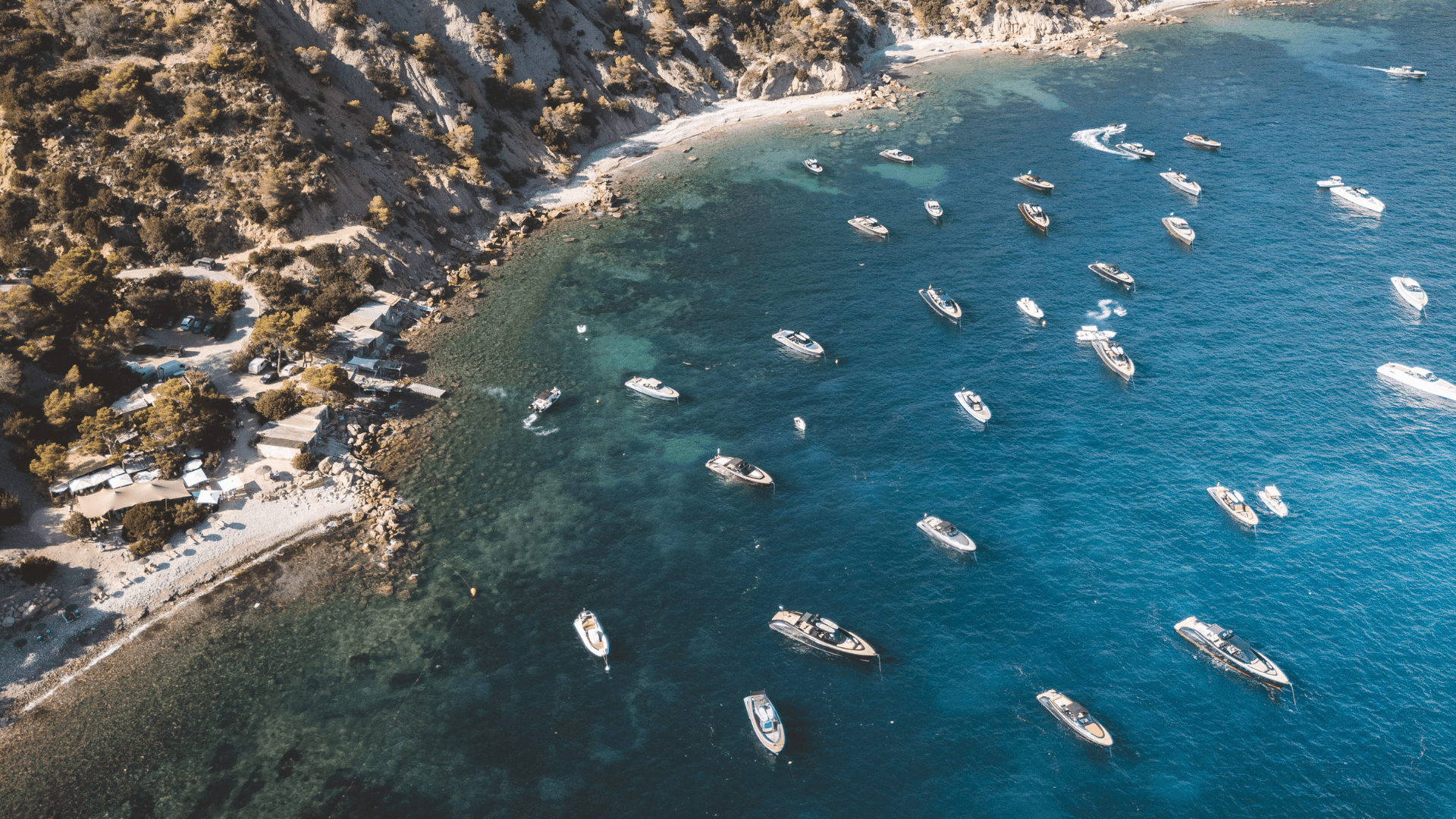 Aerial view of a rocky coastline with turquoise waters and numerous boats moored near the shore, surrounded by lush vegetation and several small structures.