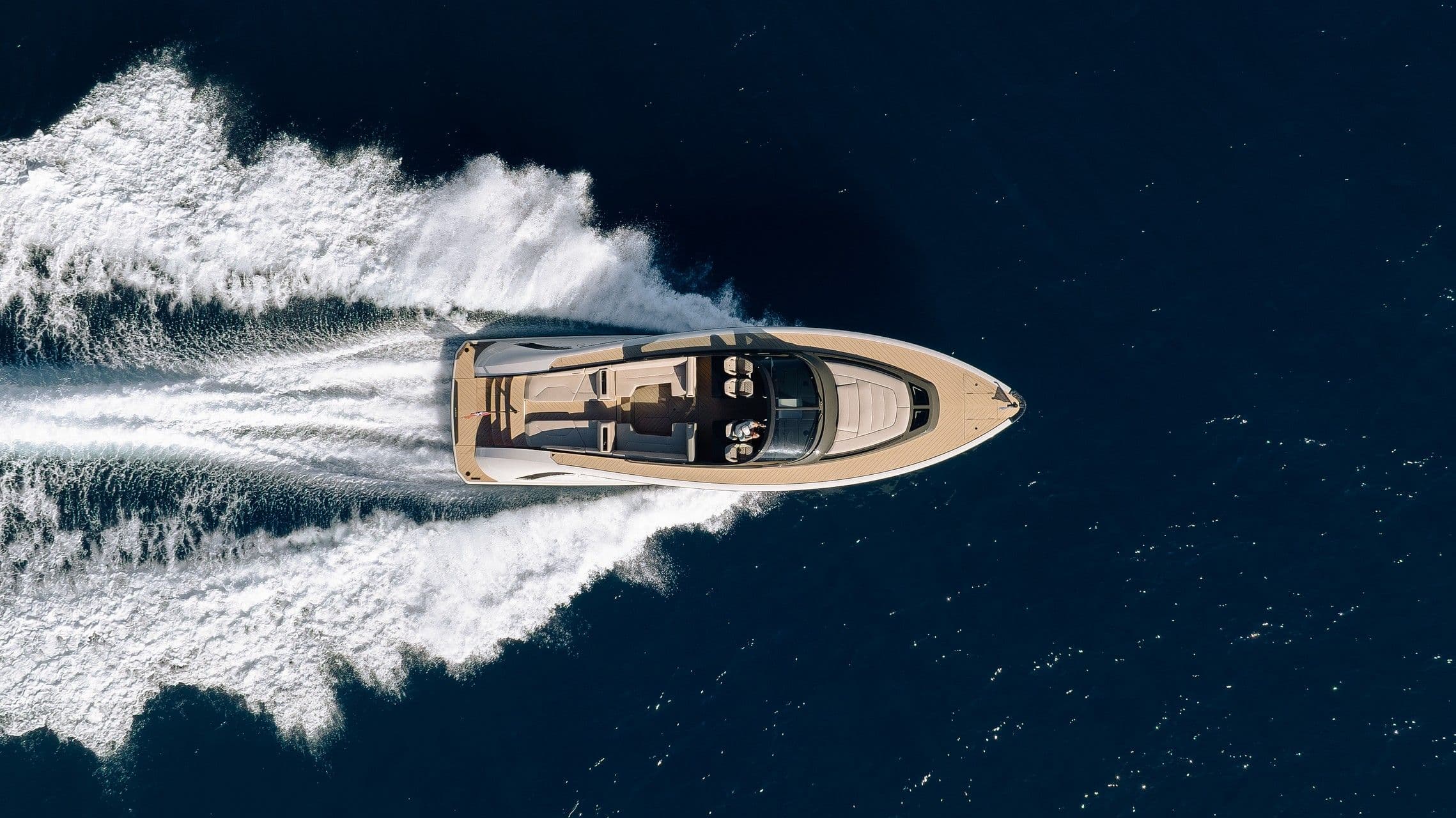Top view of a luxury motorboat cruising through deep blue water, leaving a trail of white wake behind in a sunny setting.