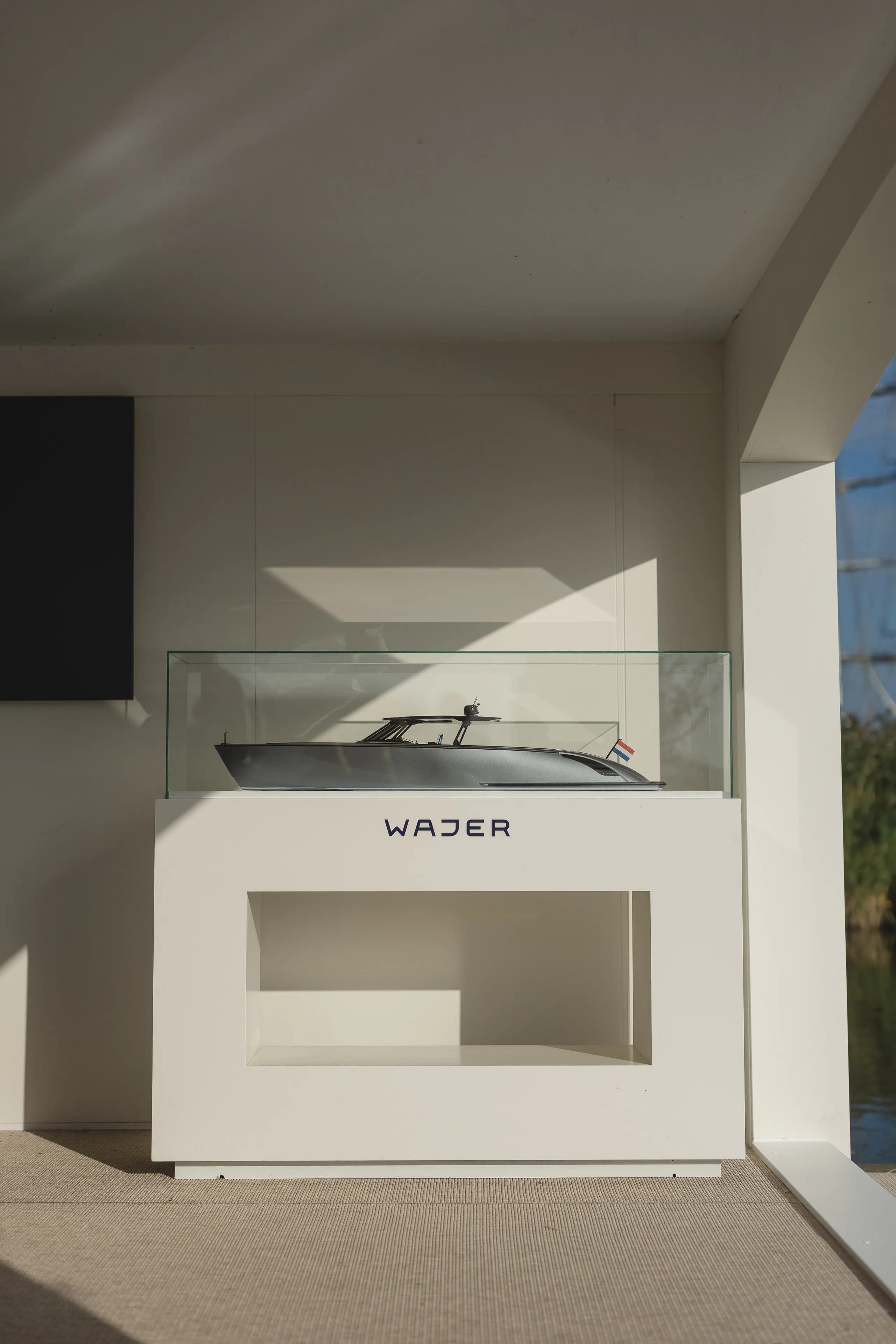 Model yacht displayed in a glass case on a white stand with "WAJER" written on it, inside a sunlit room.