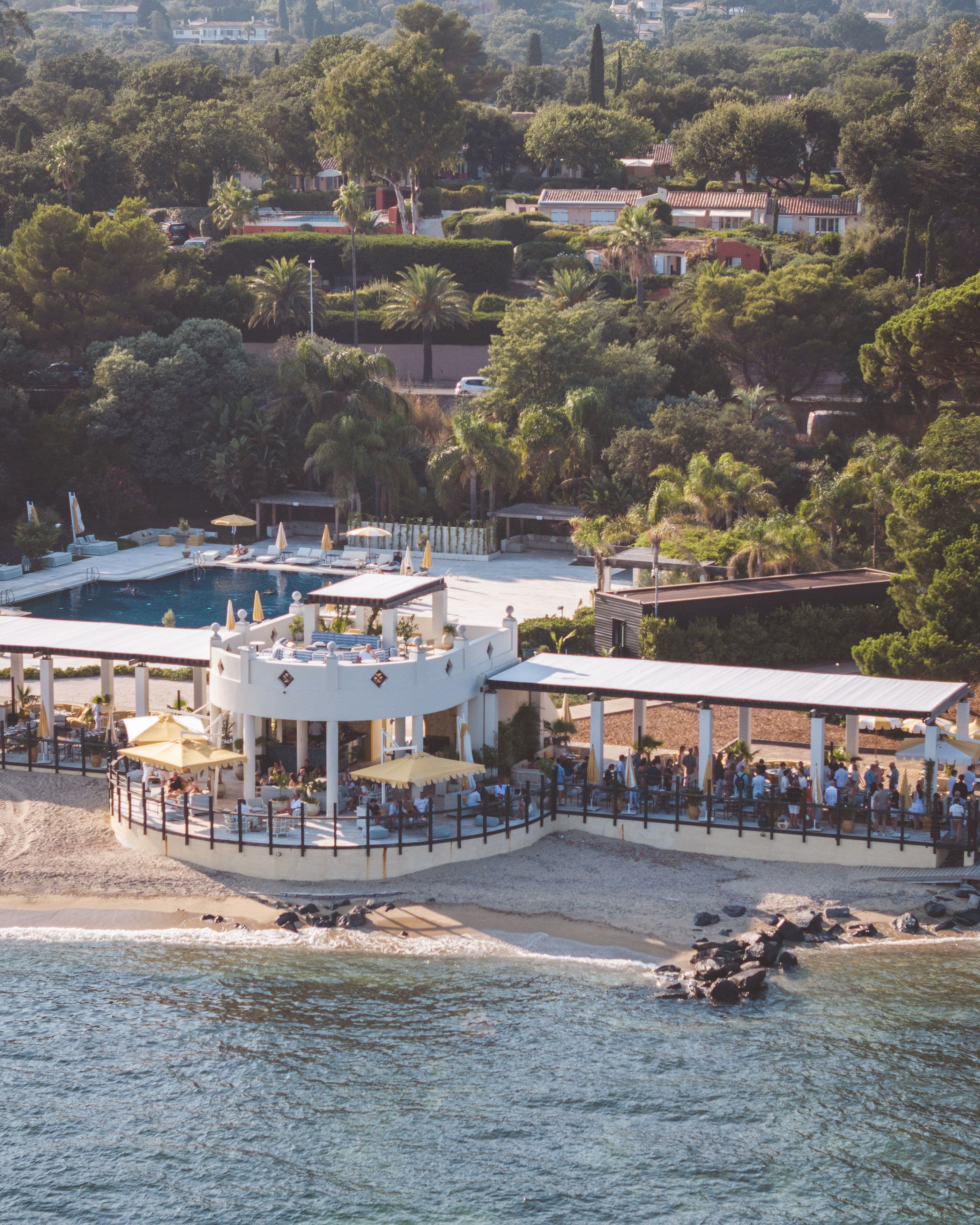 Beachfront restaurant with white circular architecture, surrounded by trees, overlooking a pool and sea. Crowds of people gather on terraces.