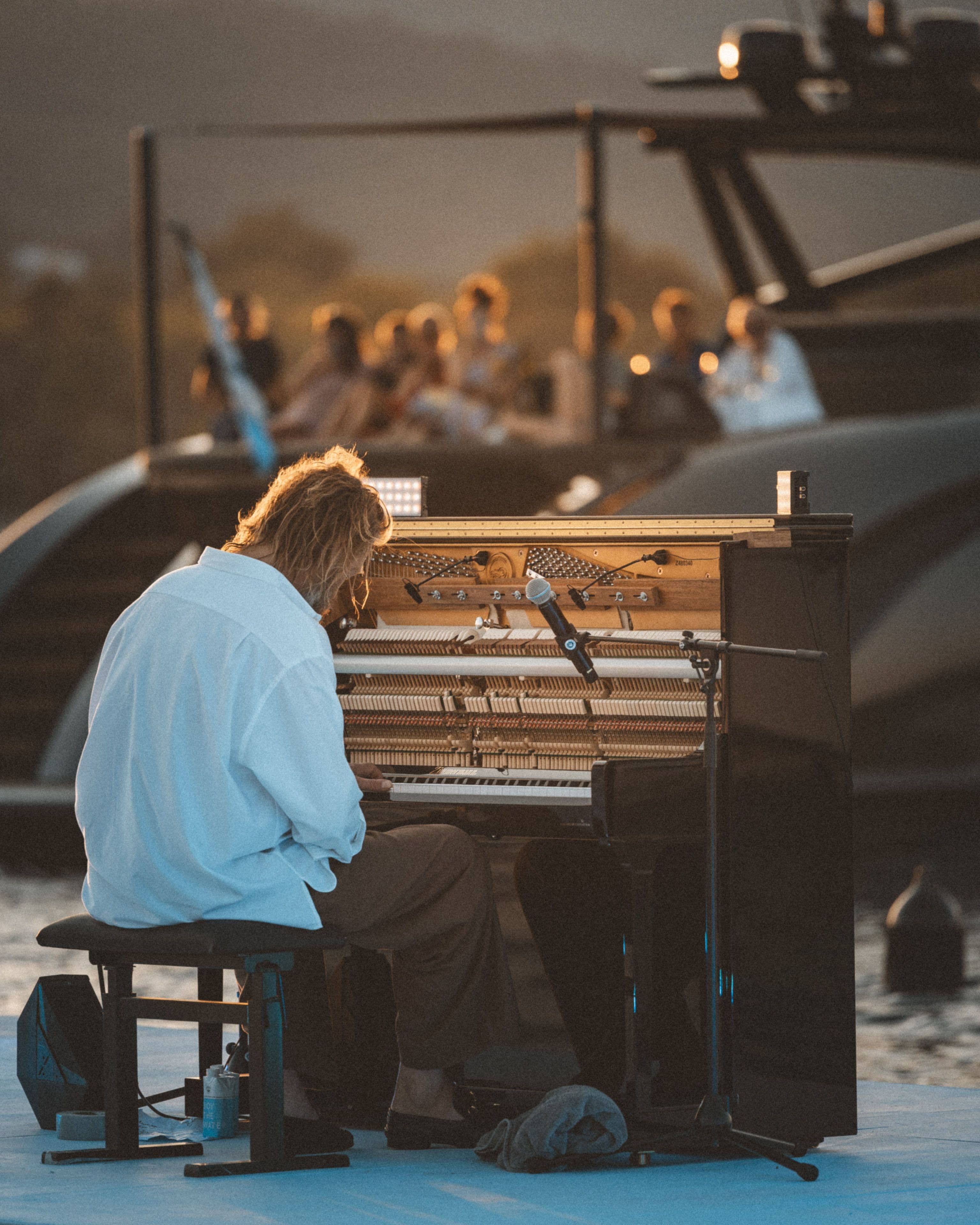 A person plays a piano outdoors with an open top, a microphone nearby, and a group of people in the background on a boat during sunset.