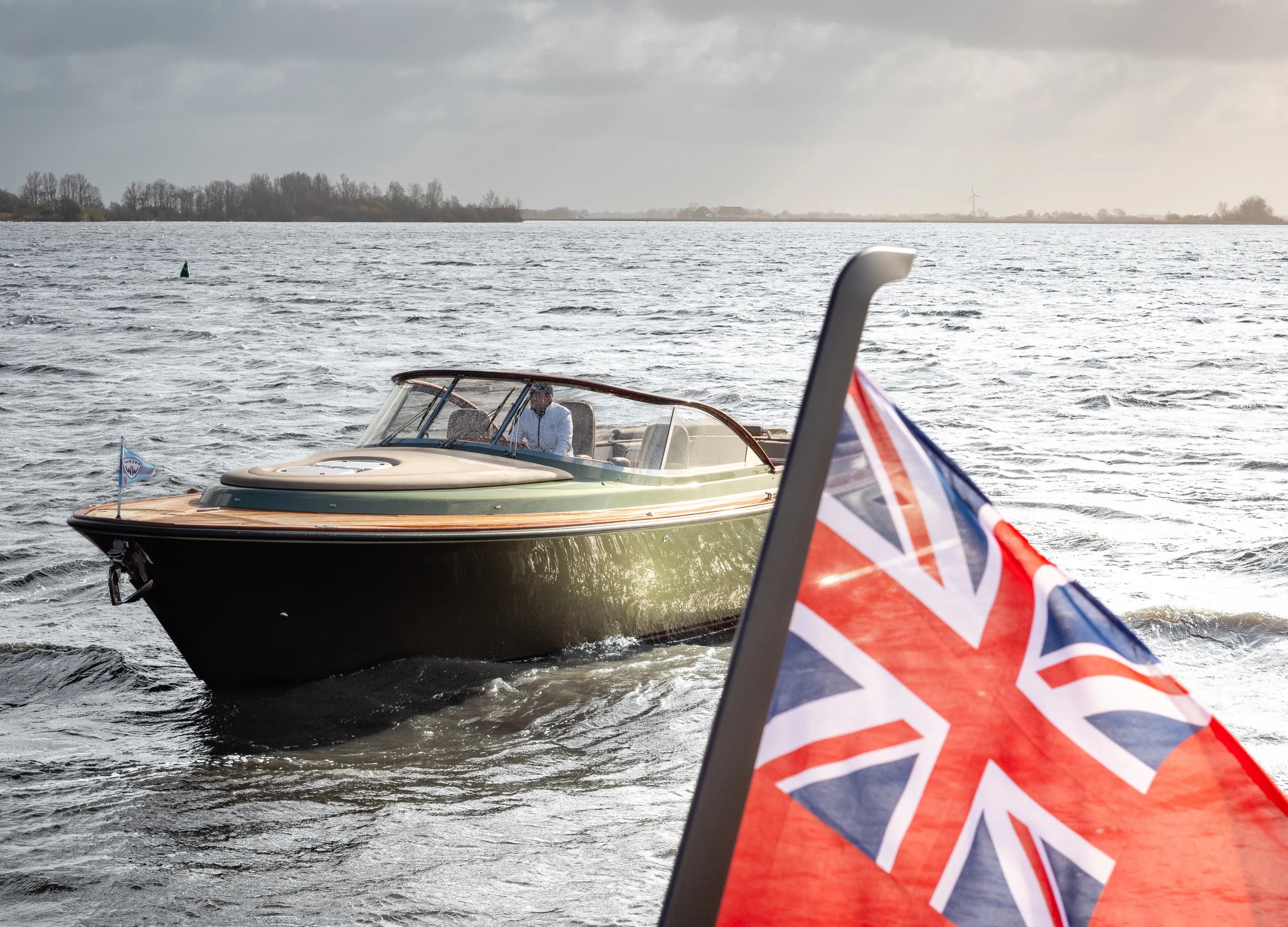 A motorboat cruising on a body of water under a cloudy sky, with a blurred Union Jack flag in the foreground.
