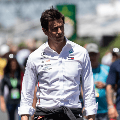 Man in a white Mercedes-branded shirt with a jacket tied around his waist, walking outdoors, surrounded by people.