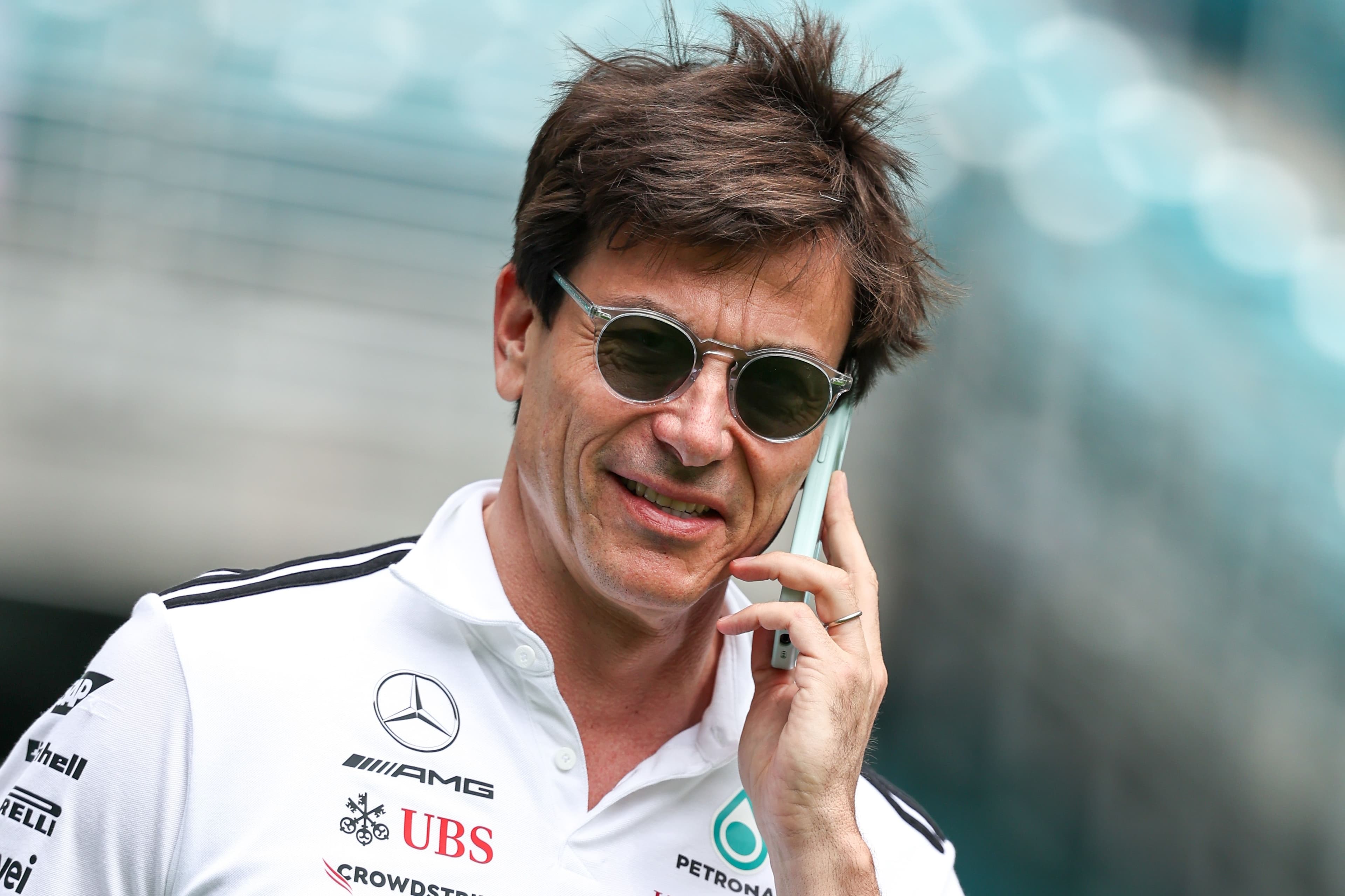 Person in a white Mercedes Petronas shirt, sunglasses, speaking on a phone outdoors.
