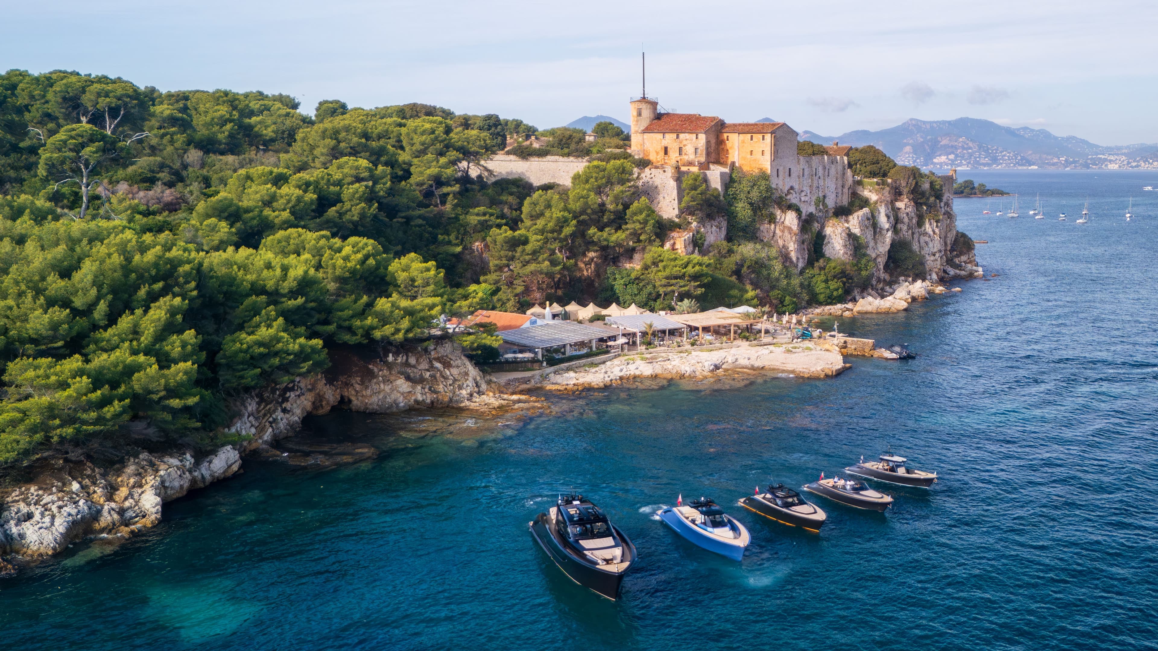 A scenic view of boats near a rocky coastline with lush greenery, a historic building on the cliff, and distant hills under a clear sky.