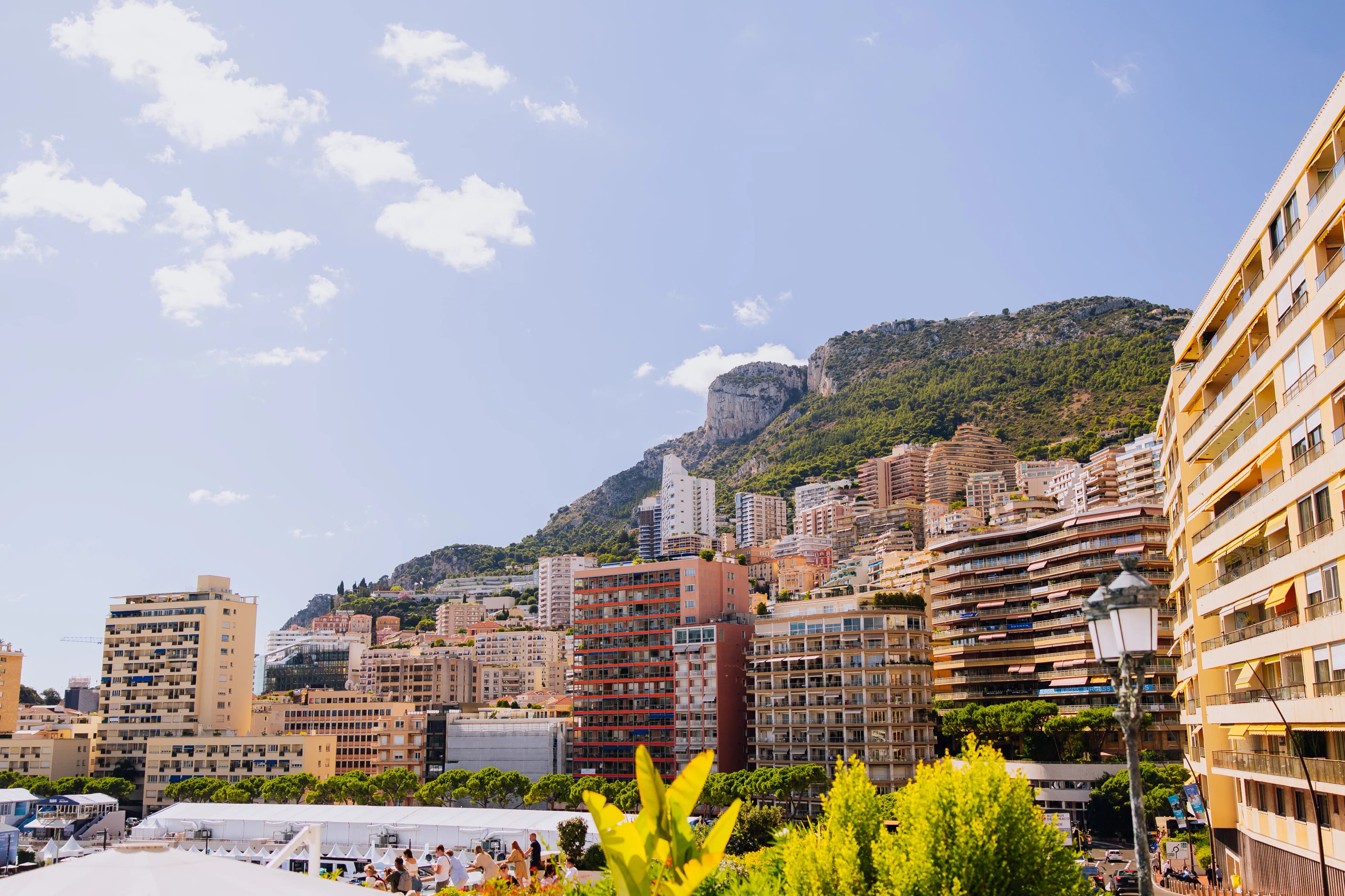 Cityscape with densely packed modern buildings, lush green hills, and a bright sky in the background. A lamppost is visible in the foreground.