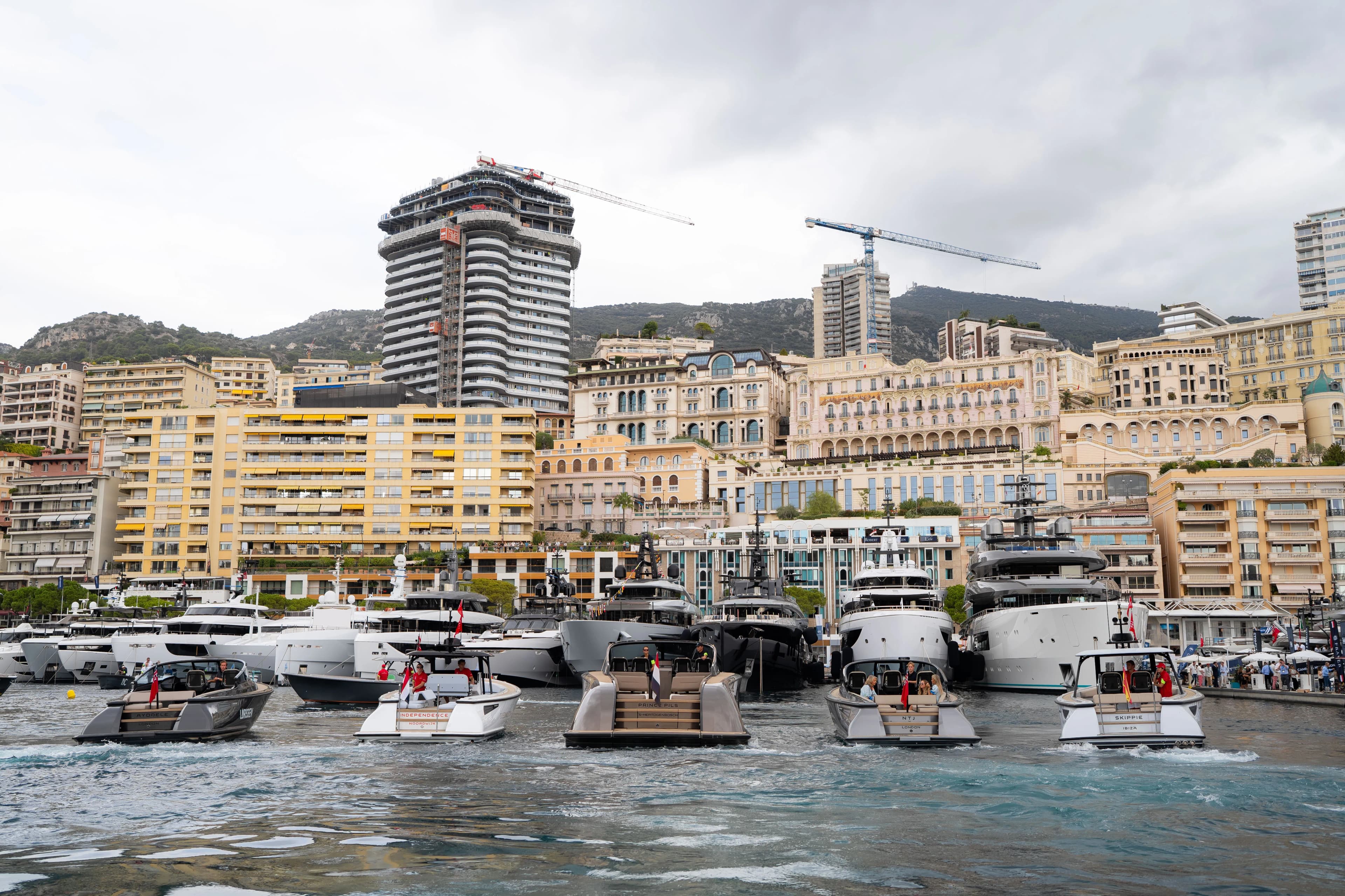 A row of yachts docked at a harbor with modern high-rise buildings and cranes in the background under a cloudy sky.