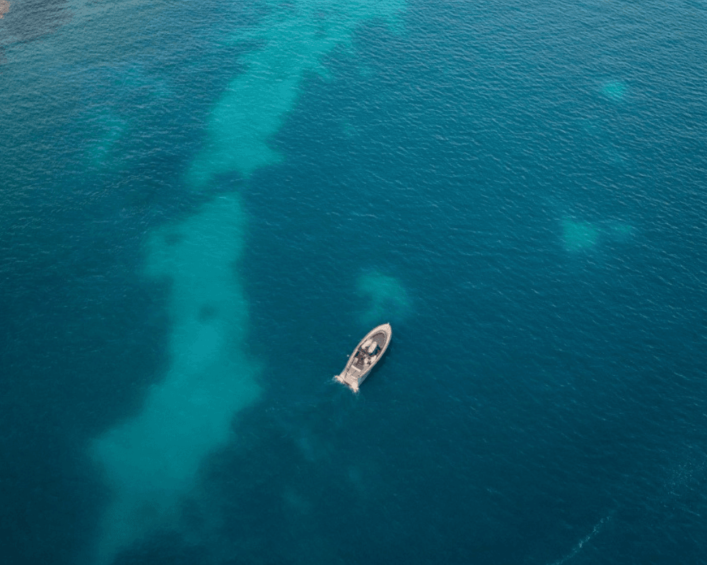 A small boat navigates through clear, deep blue ocean waters, leaving a wake behind. The sea's varying shades of blue suggest different depths.