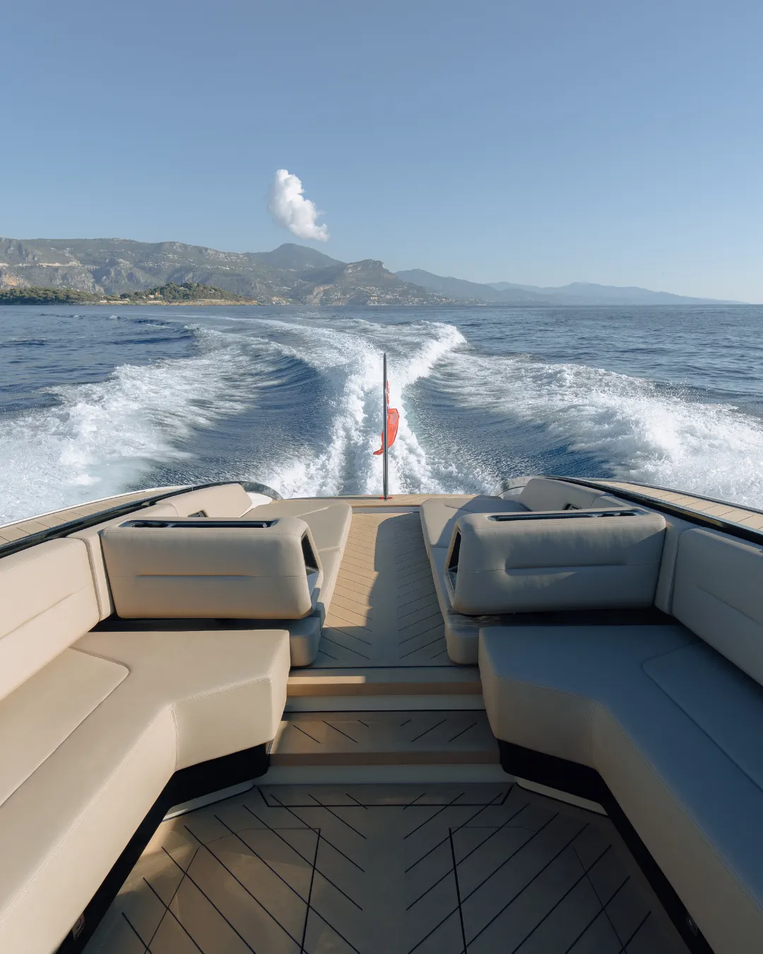 View from a boat's stern with beige seating, water wake trailing behind, and mountainous coastline under a clear blue sky.