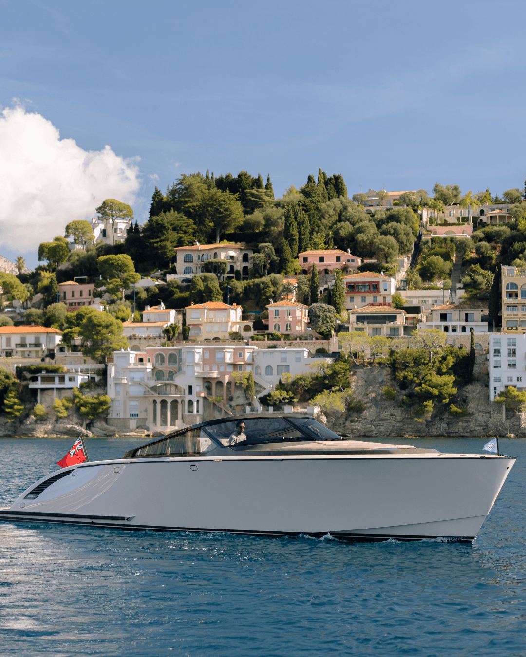 A luxury yacht cruising past Mediterranean hillside villas with lush greenery under a clear blue sky.