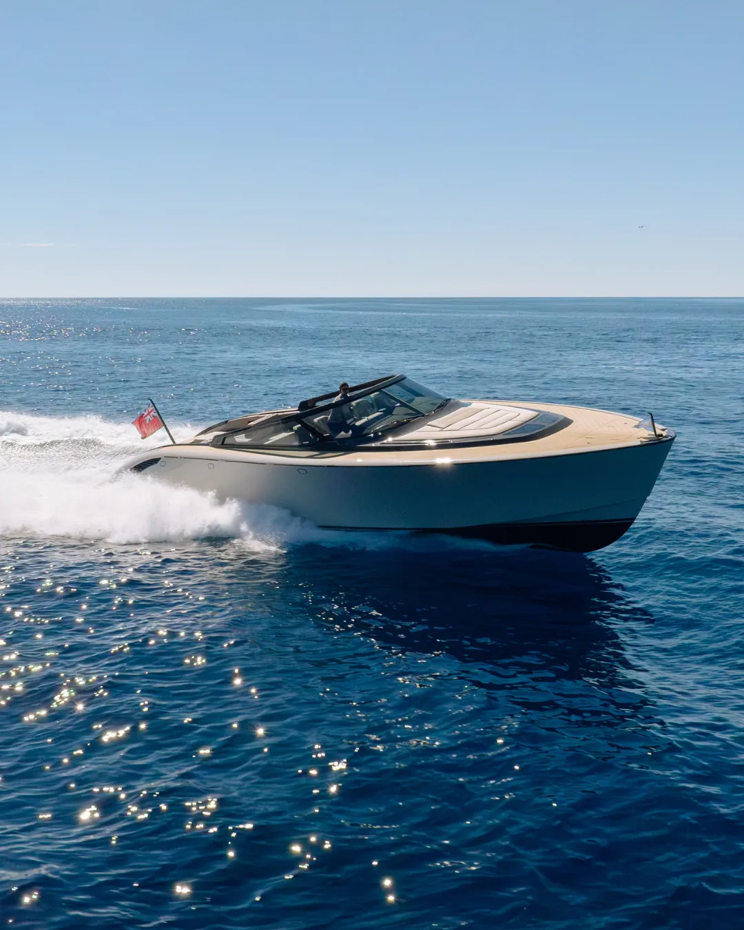 A sleek motorboat with a flag speeds through calm, sunlit ocean waters under a clear blue sky.