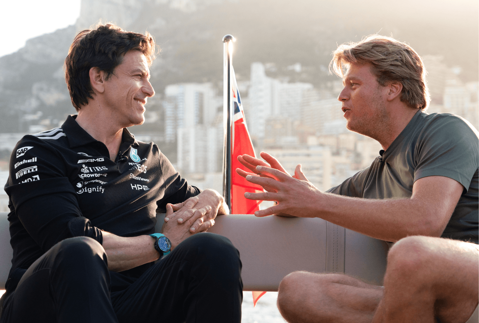 Two men seated, engaged in a lively conversation on a boat, with buildings and a flag in the background.