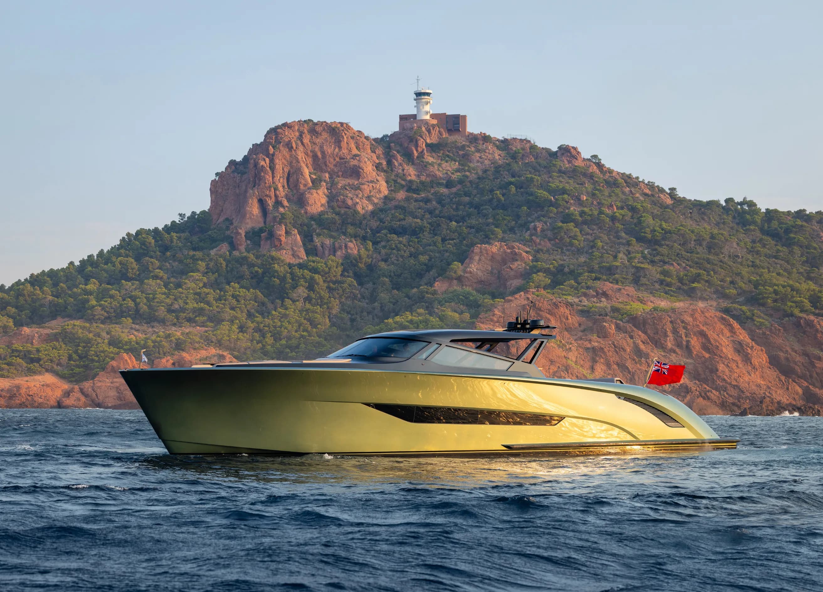 A sleek, modern yacht with a red flag sails near a rocky, tree-covered hill under a clear blue sky.