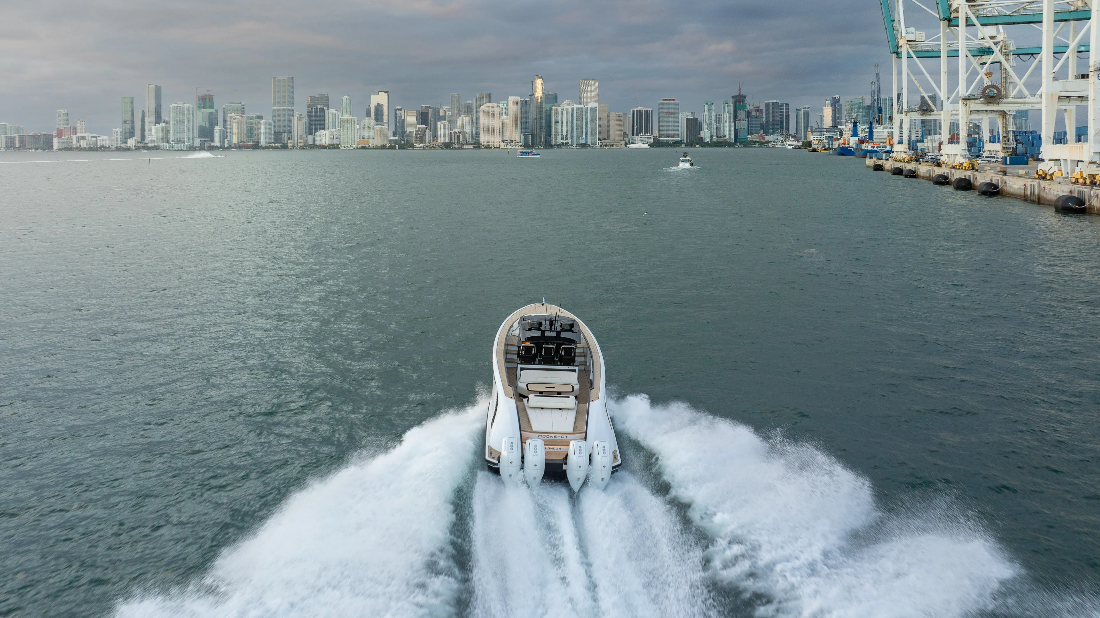 A boat cruising on water, leaving a trail of waves behind, with a city skyline and a dock with cranes in the background.