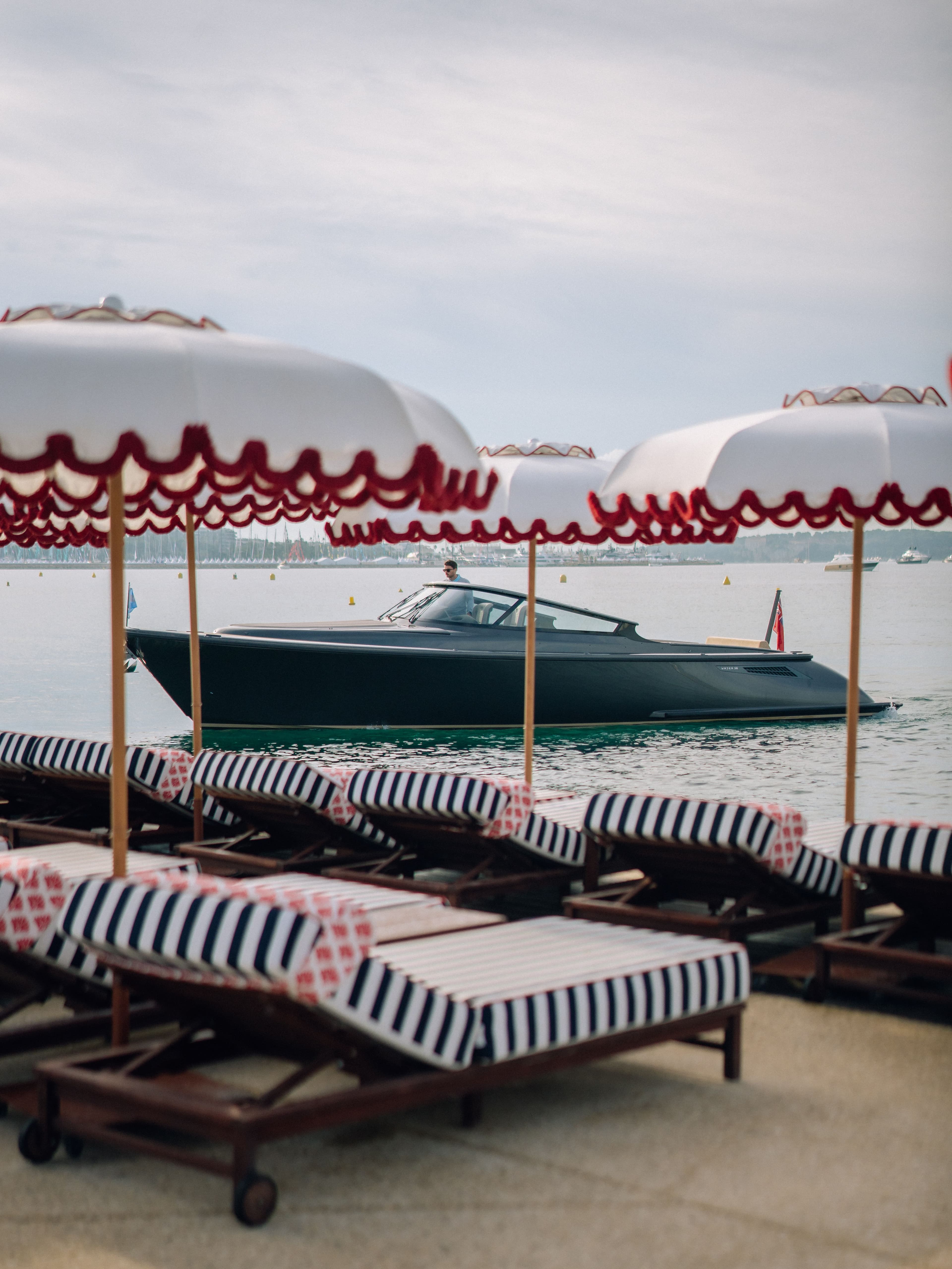 Striped lounge chairs and umbrellas on a beach overlook a sleek black boat on calm water under a cloudy sky.