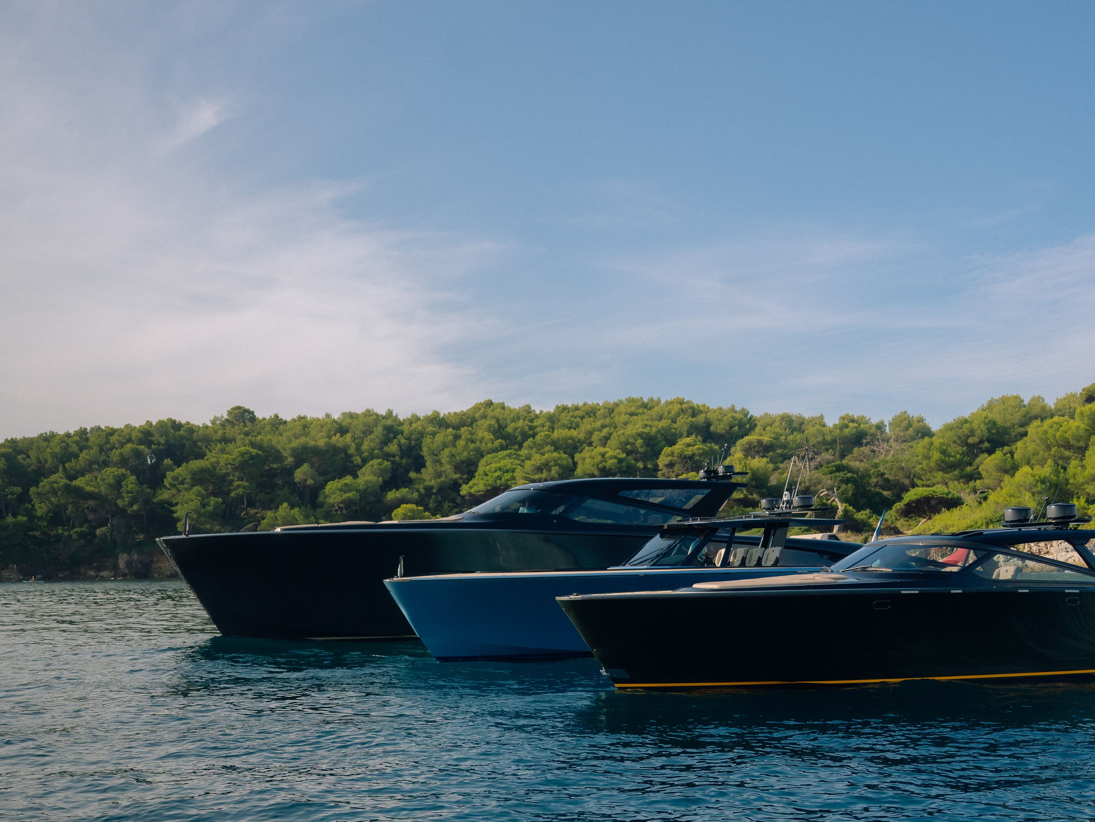 Three sleek yachts moored side by side in calm blue waters, with a lush green forest visible in the background under a clear blue sky.