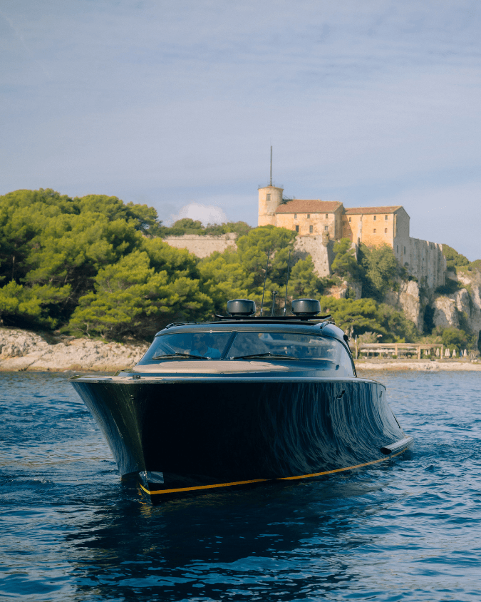A sleek black motorboat cruising on water with a historic stone castle and lush greenery in the background under a clear blue sky.